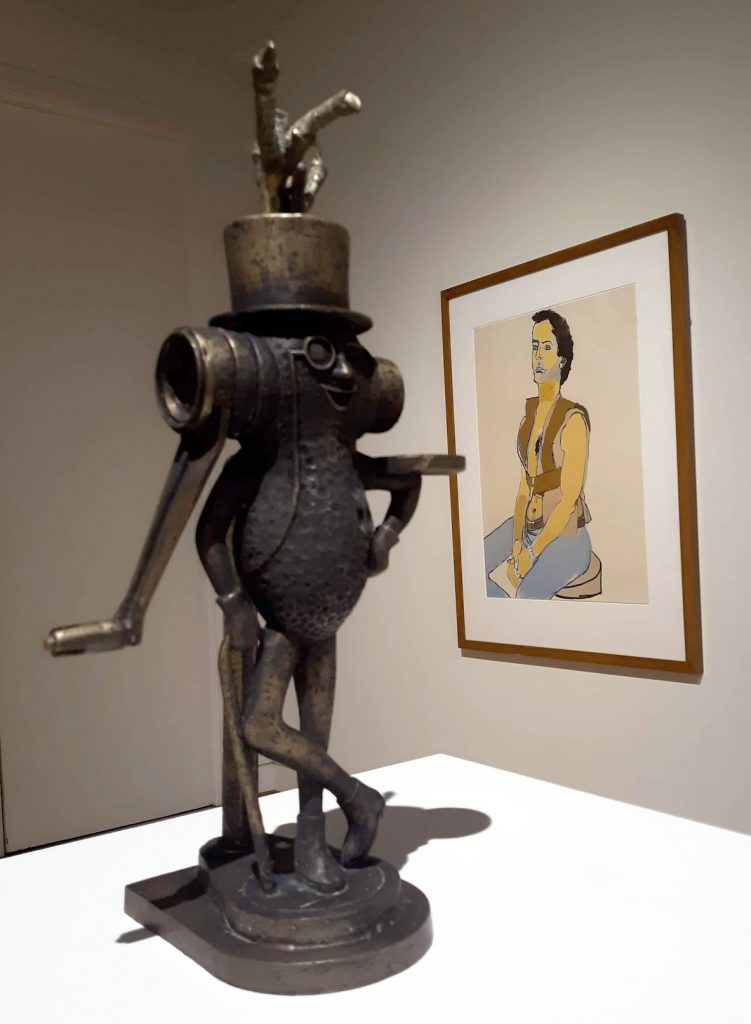 Foreground: Frank Fleming, "Mr. Peanut with Golden Egg," n.d., cast bronze (lost wax), Gift of Morgan Estate; background: Alice Neel, "Man in Harness," 1980, color lithograph, Gift of Matthew Mosca.