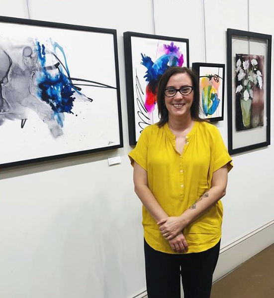 An artist poses for the camera in front of her paintings in a gallery.
