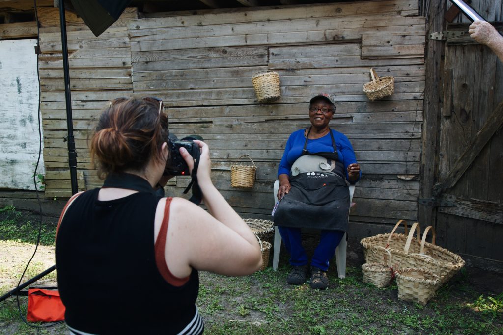 A student taking a photo of a basket weaver sitting against a workshed.